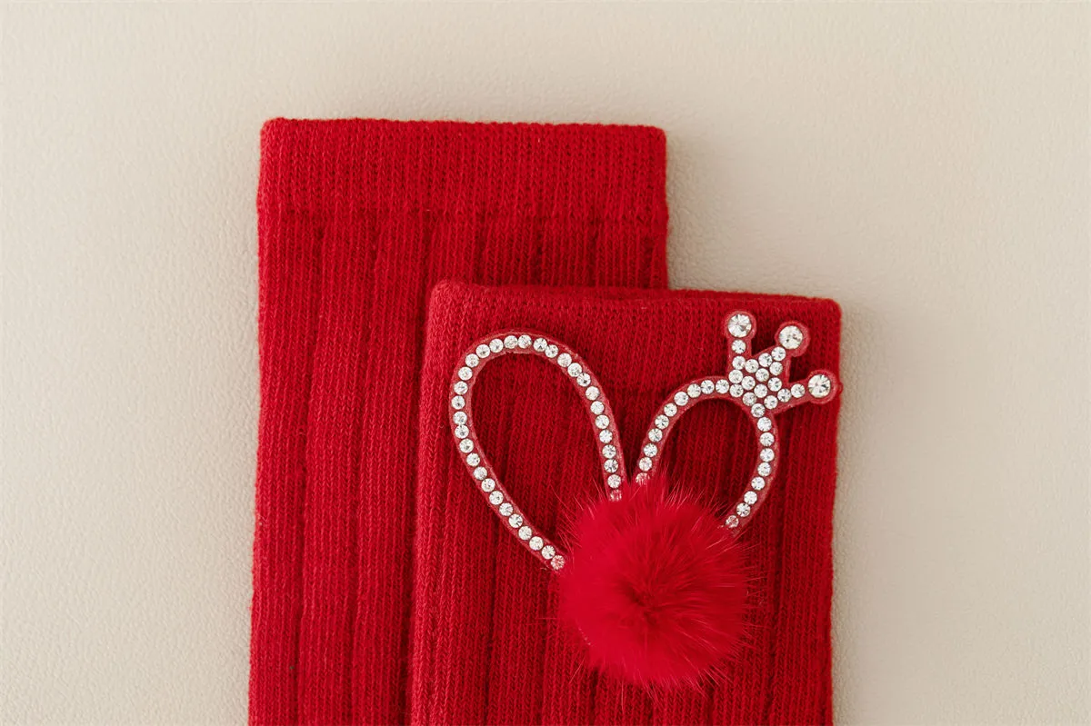Close-up of red baby socks featuring a rhinestone crown and fluffy pom pom detail. Close-up of red baby socks featuring a rhinestone crown and fluffy pom pom detail.